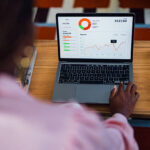A focused individual uses a laptop on a wooden desk displaying a colorful business dashboard with charts and graphs, conveying analytics, productivity, and data-driven decision making in a modern setting.